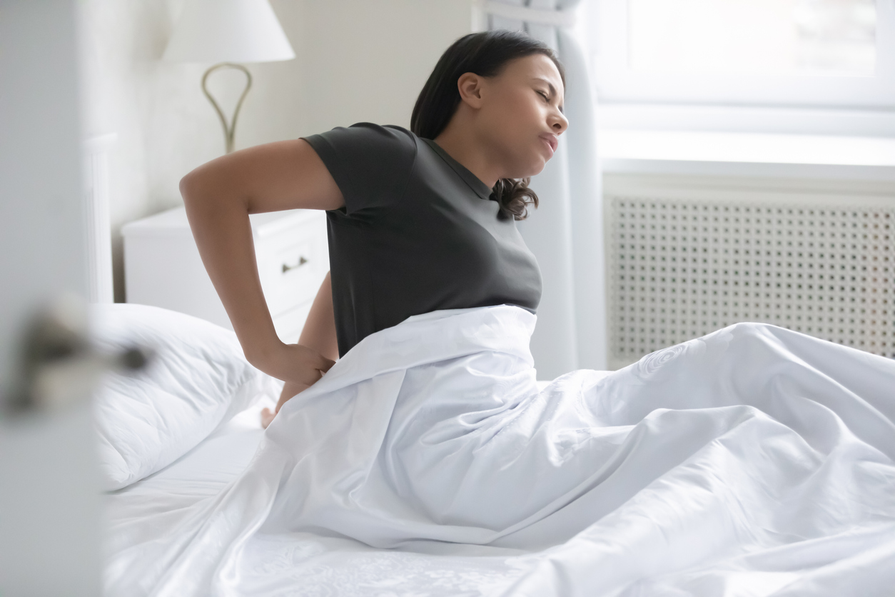 Woman holds her lower back in pain, while sitting up in bed.